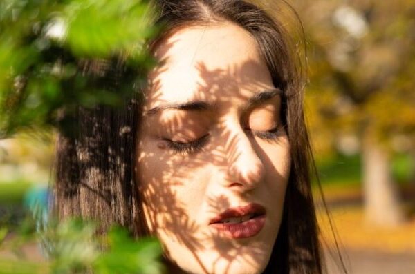 A woman with eyes closed in sunlight, leaf shadows cast on her face, creating a serene and artistic portrait.